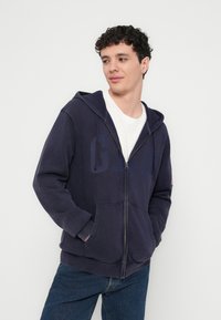 Námořnicky modrá mikina na zip s klokánkovou kapsou a kapucí se stahovací šňůrkou. Vyrobená z měkké látky, s jemným potiskem a žebrovanými manžetami.