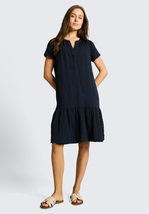 Jonge vrouw draagt een zwarte, gestructureerde jurk tot op de knie met korte mouwen en beige instapsandalen, staand met handen achter de rug.