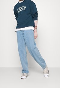 Modrá mikina Levi's s bílým logem, oblečená přes bílou košili, spárovaná se světle modrými volnými džínami a šedými teniskami.