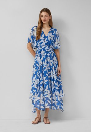 Vrouw draagt een midi-jurk met blauwe en witte bloemenprint, pofmouwen en sandalen, staand tegen een effen grijze achtergrond.
