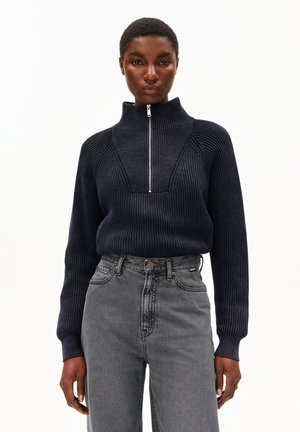 Vrouw in een marineblauwe geribbelde trui met een rits aan de voorkant en hoge kraag, gecombineerd met hoge grijze jeans, staand tegen een effen achtergrond.