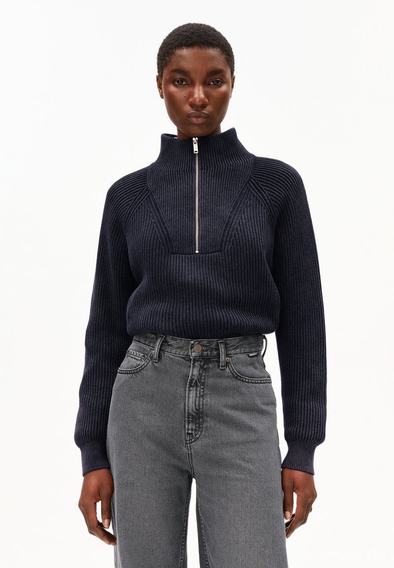 Frau, die einen marineblauen gerippten Pullover mit einem Reißverschluss an der Vorderseite und einem hohen Kragen trägt, kombiniert mit hochgeschnittenen grauen Jeans, stehend vor einem schlichten Hintergrund.