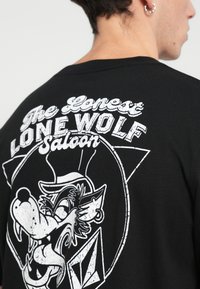 T-shirt preta com uma impressão gráfica branca de um lobo a usar um chapéu, com o texto "O Saloon do Lobo Solitário" e acentos decorativos.