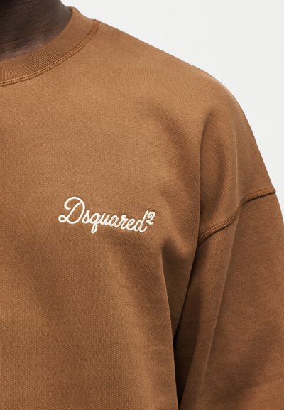 Καφέ φούτερ με λευκό κεντημένο λογότυπο "Dsquared²" στο αριστερό στήθος, που φοριέται από ένα άτομο με σκουρόχρωμο δέρμα.