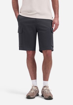 Man draagt donkergrijze cargoshorts, witte sokken, beige sneakers en een lichtgekleurde shirt, staand met handen gedeeltelijk in de zakken.