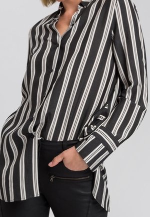 Person wearing a black and white vertically striped button-up shirt with one hand in the pocket of black pants with a zipper detail.