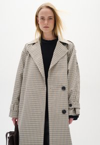 Cappotto trench doppiopetto in un motivo a scacchi beige e nero, con un ampio colletto, dettagli con bottoni e una vestibilità rilassata.