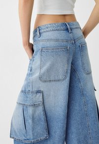 Vaaleansiniset farkkucargo-housut, joissa on kaksi suurta sivutaskua, suora lahje ja haalistunut pintastruktuuri näkyvine ompelutietoineen.