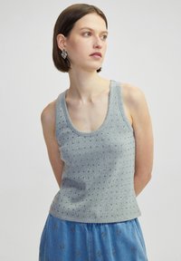 Grijze gebreide tanktop met kleine decoratieve studs, brede halslijn en een getailleerd silhouet. Gecombineerd met een blauwe patroonrok.
