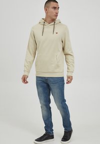 Felpa cappuccio beige con tasca frontale, caratterizzata da lacci decorati e un piccolo logo sul petto, abbinata a jeans blu e sneakers nere.