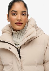 Beige pufferjack met een hoge kraag, ritsluiting en drukknoopjes. Draagt over een gemêleerde gebreide trui in crème en meerdere kleuren.