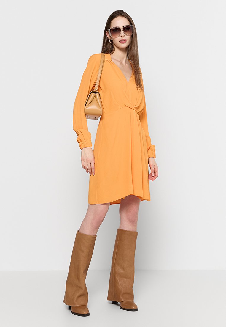 Pomarańczowa sukienka w stylu wrap z długimi rękawami, z węzłem w talii i rozszerzanym dołem. Połączona z brązowymi botkami do kolan i beżową torbą.