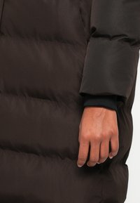 Cappotto puffer marrone con design trapuntato, che presenta una finitura testurizzata, polsini elastici e una manica aderente vicino al polso.