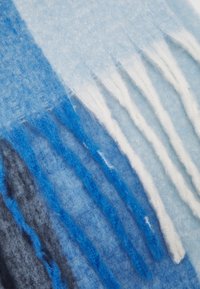 Écharpe en laine avec des teintes de bleu, noir et blanc, dotée d'une finition douce et texturée. Comprend un détail de franges le long d'un bord.