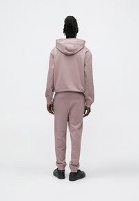 Mikina s kapucňou a zodpovedajúce tepláky z jemnej ružovej látky. Má rebrované manžety a pás, nosené s čiernymi topánkami. Pohľad zozadu.