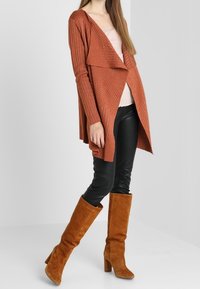 Rostfarbener Strickcardigan mit offenem Frontbereich, strukturiertem Stoff, geschichteter Gestaltung und langen Ärmeln. Kombiniert mit schwarzen Lederleggings und hellbraunen kniehohen Stiefeln.