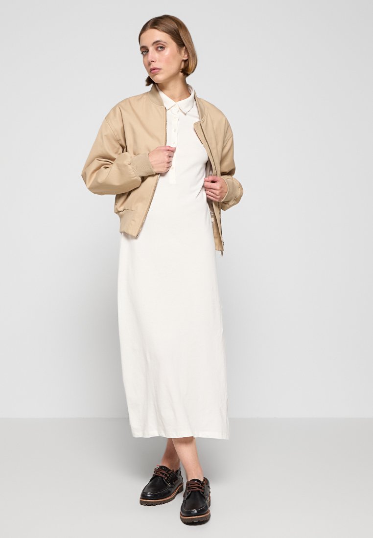 Beige bomberjack over een witte jurk tot de knie, gecombineerd met zwarte veterschoenen. Glad materiaal met minimale details en een eenvoudig ontwerp.
