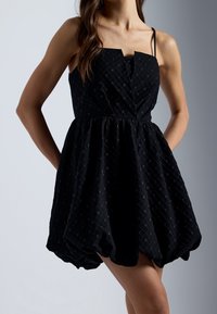 Robe noire sans manches avec un corsage ajusté, jupe évasée, tissu texturé arborant un motif en diamant, et poches sur les côtés.