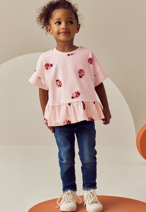 Ung pige med krøllet hår iført en pink top med røde mariehøner, blå jeans og hvide sneakers, der står på en rund orange platform.
