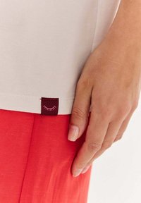 Ruka opřená o červené kalhoty, oblečená v béžovém tričku s malou bordó nášivkou s designem růžových řas.