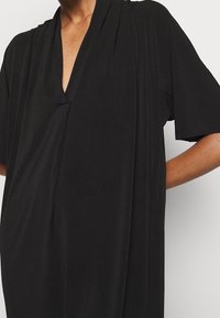 Svart klänning med kort ärm och djup V-ringning samt draperad design. Mjuk, slät tygkvalitet med minimal sömdetaljering för en avslappnad passform.