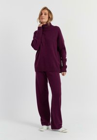 Overmaatse paarse gebreide trui met een hoge col en brede mouwen, gecombineerd met losse paarse broek en witte sneakers.