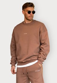 Brown cotton sweatshirt with ribbed cuffs and neckline, featuring a small embroidered logo. Paired with matching trousers and sunglasses.