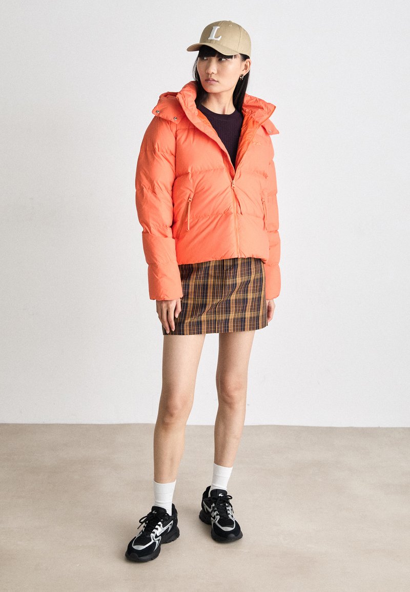 Orangene Pufferjacke mit Kapuze, Reißverschluss vorne und zwei Taschen; kombiniert mit einem karierten Rock, weißen Socken und schwarzen Sneakers.