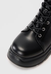 Μαύρο δερμάτινο μποτάκι με κορδόνια, με λείο στρογγυλό δάχτυλο και παχύ καουτσούκ πάτο σε λευκό φόντο.