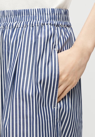 Main dans la poche d’un pantalon à taille élastique rayé vertical bleu et blanc, montrant la texture du tissu et le détail de la poche.