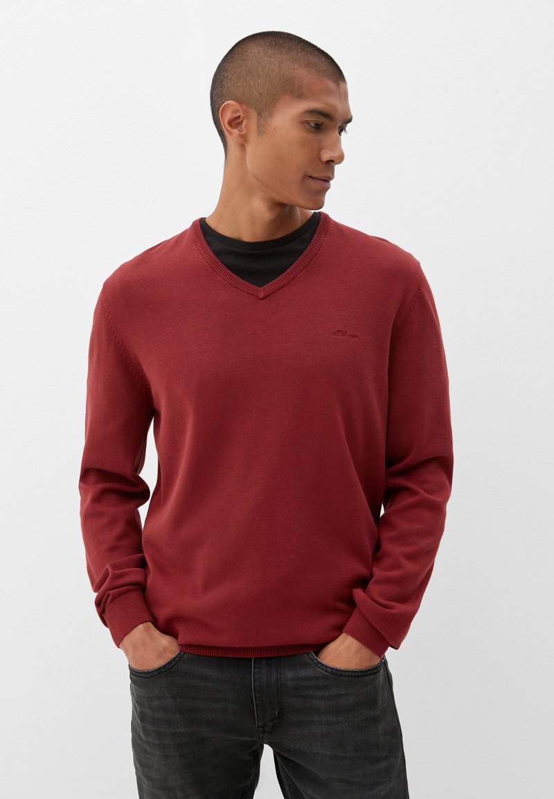 Rode v-hals trui met lange mouwen, gebreide textuur, geribbelde manchetten en een subtiel logo-detail. Draagt over een zwart t-shirt en gecombineerd met donkere jeans.