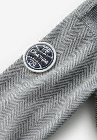 Szara tkanina w wzór herringbone posiada okrągłą wyszywaną naszywkę z niebieskimi i białymi akcentami, na której znajduje się napis "Champ '09".