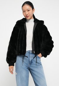 Sort faux fur bomberjakke med lynlås foran og elastiske ærmer. Har snore i kanten og en blød, struktureret finish.