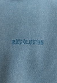 Lichtblauwe stof met een ribbelstructuur met daarop het geborduurde woord "REVOLUTIE" in donkerder blauw.
