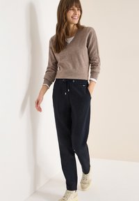 Pull col en V marron clair associé à un pantalon bleu marine foncé et fuselé. La tenue comprend une taille à cordon de serrage et des baskets de couleur claire.