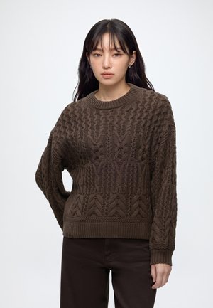 Young woman with long dark hair wears dark brown textured knit sweater and dark pants, standing against plain light background.