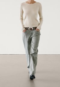 Beige ribbed long-sleeve top paired with light gray straight-leg jeans, featuring a brown belt with a circular silver buckle. Black shoes.