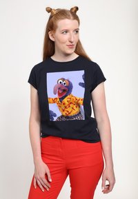 Svart kortärmad t-shirt med en färgglad grafik av en karaktär i en gul skjorta med röda och orange mönster. Röda tajta byxor.