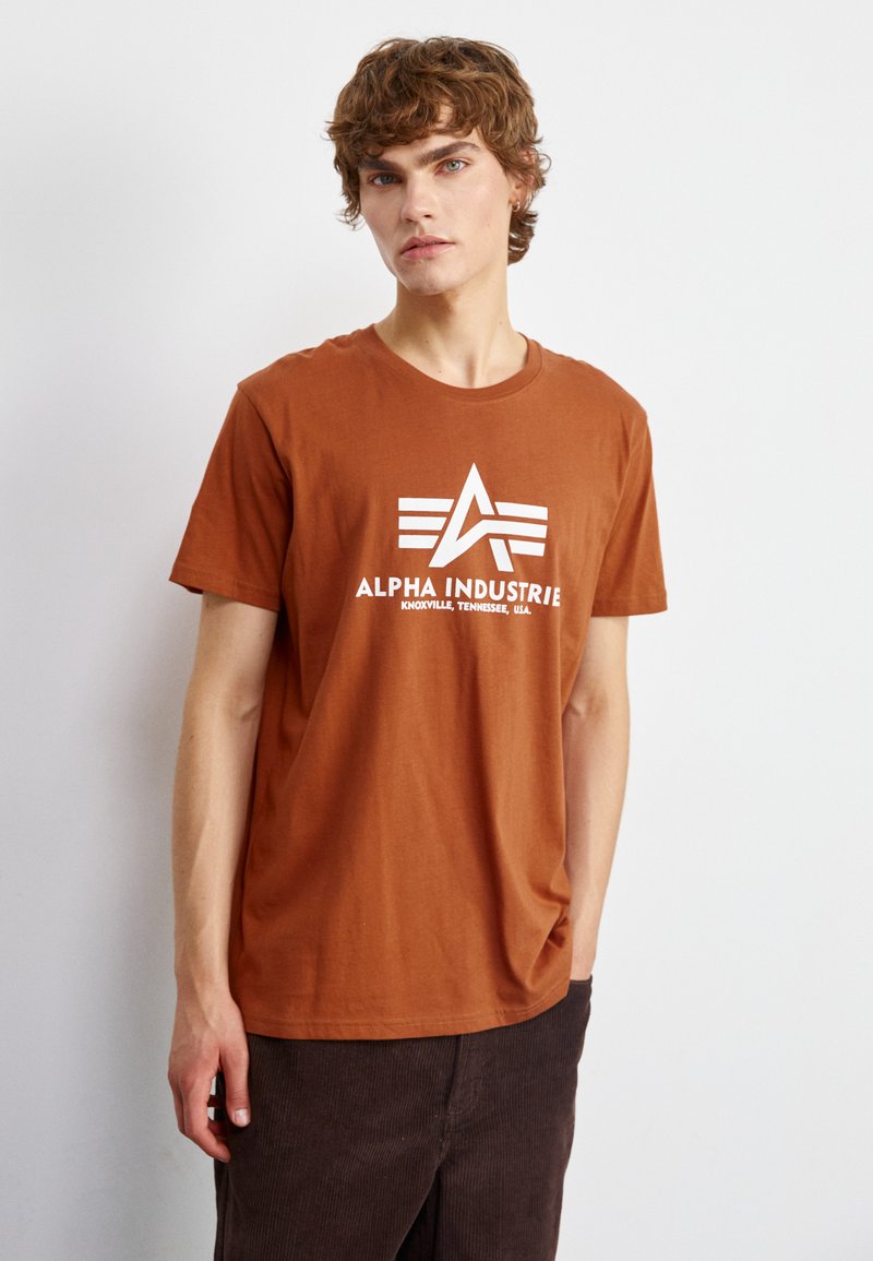 Camiseta de algodón marrón con un estampado de logo blanco que dice "ALPHA INDUSTRIE" y tres rayas paralelas. Mangas cortas y cuello redondo.