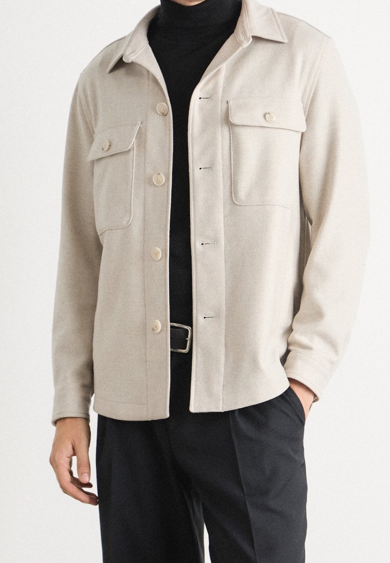 Chaqueta de mezcla de lana beige con cuello de camisa, bolsillos en el pecho y cierre de botones, combinada con un cuello de tortuga negro y pantalones oscuros.