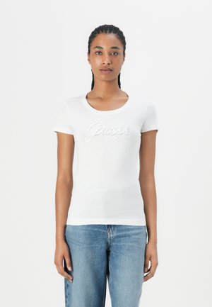 T-shirt blanc à manches courtes avec un col rond, présentant un logo "Guess" en strass sur la poitrine. Tissu doux et lisse.