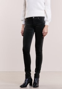 Žena oblečená v černých slim-fit kalhotách, černých kotníkových botách na block podpate, a bílém dlouhém svetru s chloupky, stojící na hladké podlaze.