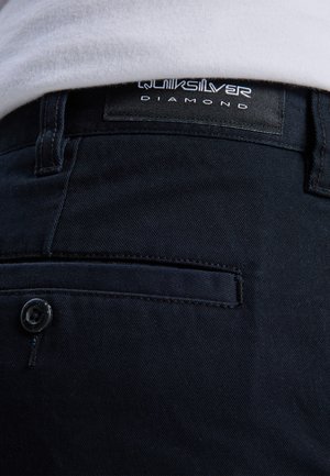 Donkerblauwe broek met een gestructureerde stof, een achterzak, een knoopsluiting en een zwart label met de tekst "Quiksilver Diamond."