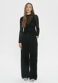 Top negro transparente de manga larga con detalles de encaje, combinado con unos vaqueros negros de corte holgado. La modelo está de pie con las manos en los bolsillos, llevando zapatos plateados.