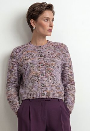 Frau mit kurzen Haaren, die silberne Creolen, eine mehrfarbige strukturierte Strickjacke mit Knöpfen und violette High-Waist-Hosen trägt, mit Händen in den Taschen.