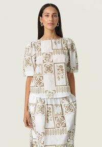 Vit blus med en strukturerad passform, puffärmar och ett repetitivt mönster av gyllene blommor. Matchas med en kjol som har samma design.