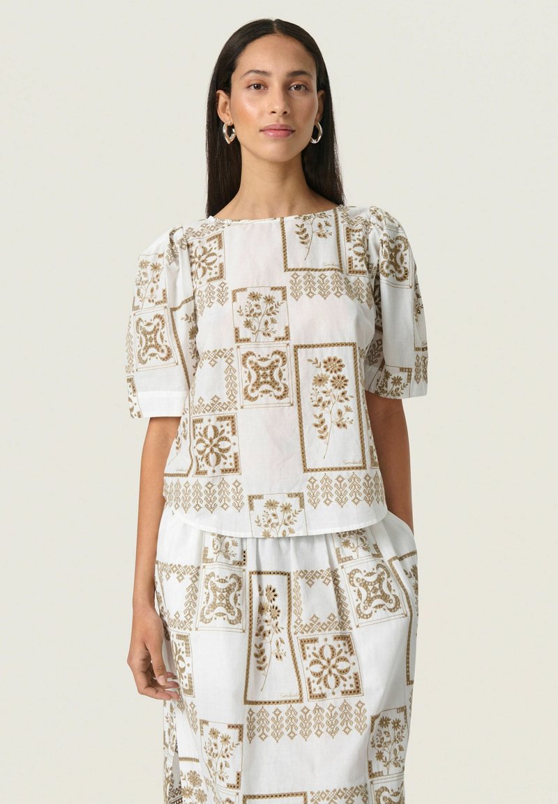 Vit blus med en strukturerad passform, puffärmar och ett repetitivt mönster av gyllene blommor. Matchas med en kjol som har samma design.