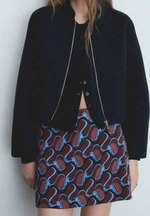 Femme portant une veste noire courte, un haut noir boutonné et une jupe marron avec des motifs abstraits bleus, debout devant un fond uni.