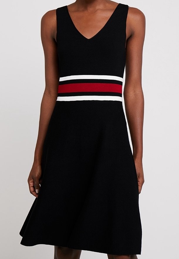 Robe noire sans manches avec un col en V et des rayures horizontales rouges, blanches et noires à la taille, portée par une personne debout devant un fond uni.