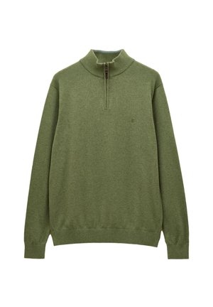 Mann trägt grünen Viertel-Reißverschluss-Pullover über kariertem Hemd und beigen Hosen, steht mit Händen in den Taschen vor einfarbigem beigem Hintergrund.